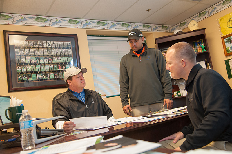 Three men in a golf course maintenance shop office talking and listening to each other