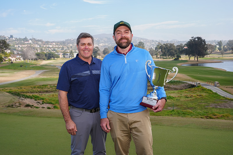 Two men standing outside. The man on the right is holding a trophy