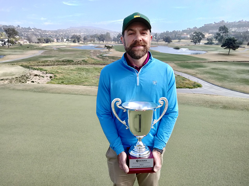 A man wearing a ball cap and blue sweater holds a trophy