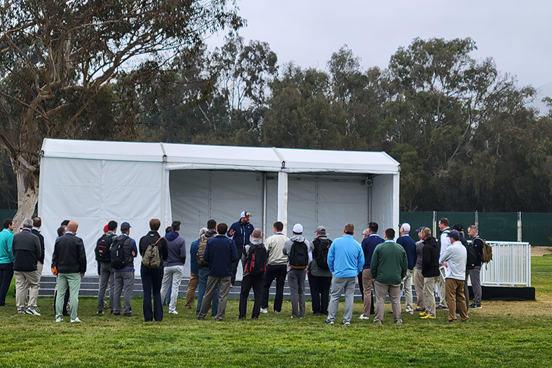 A group of men stand in front of a white tent where a man is speaking on a stage
