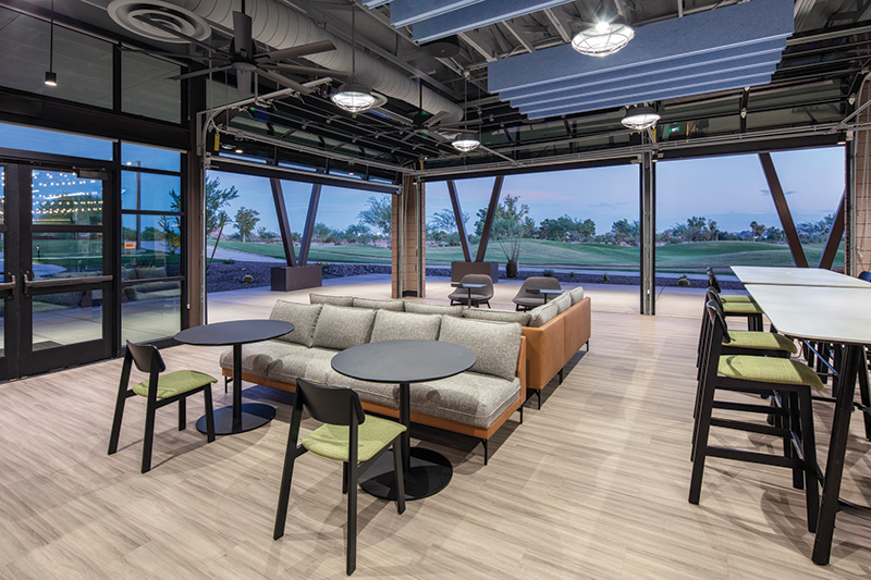 A lounge with couches, chairs, tables and a bar-height counter with stools and windows looking out onto a golf course in the early evening