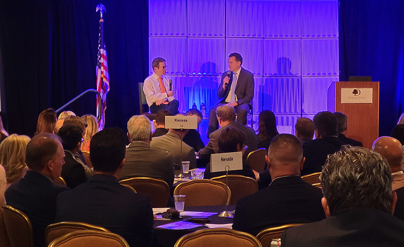 Two men sitting and talking on a stage in a conference room to a large group of people