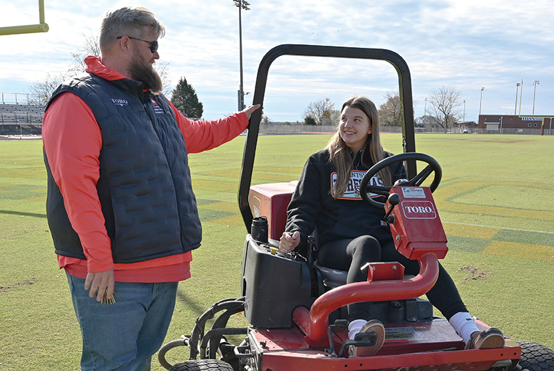 Drew Miller and Elizabeth Mirko. Mirko is sitting on a riding mower on a football field as Miller talks to her