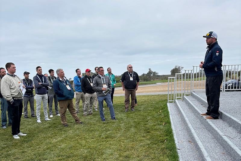 Man standing on an outdoor stage talking to a group of other men