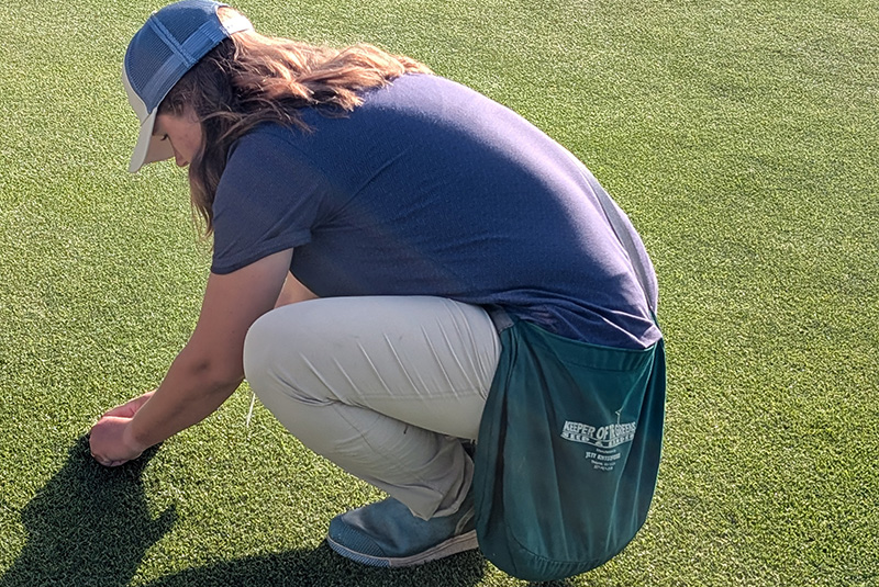 Woman with long hair wearing a baseball cap crouched on a kikuyugrass green